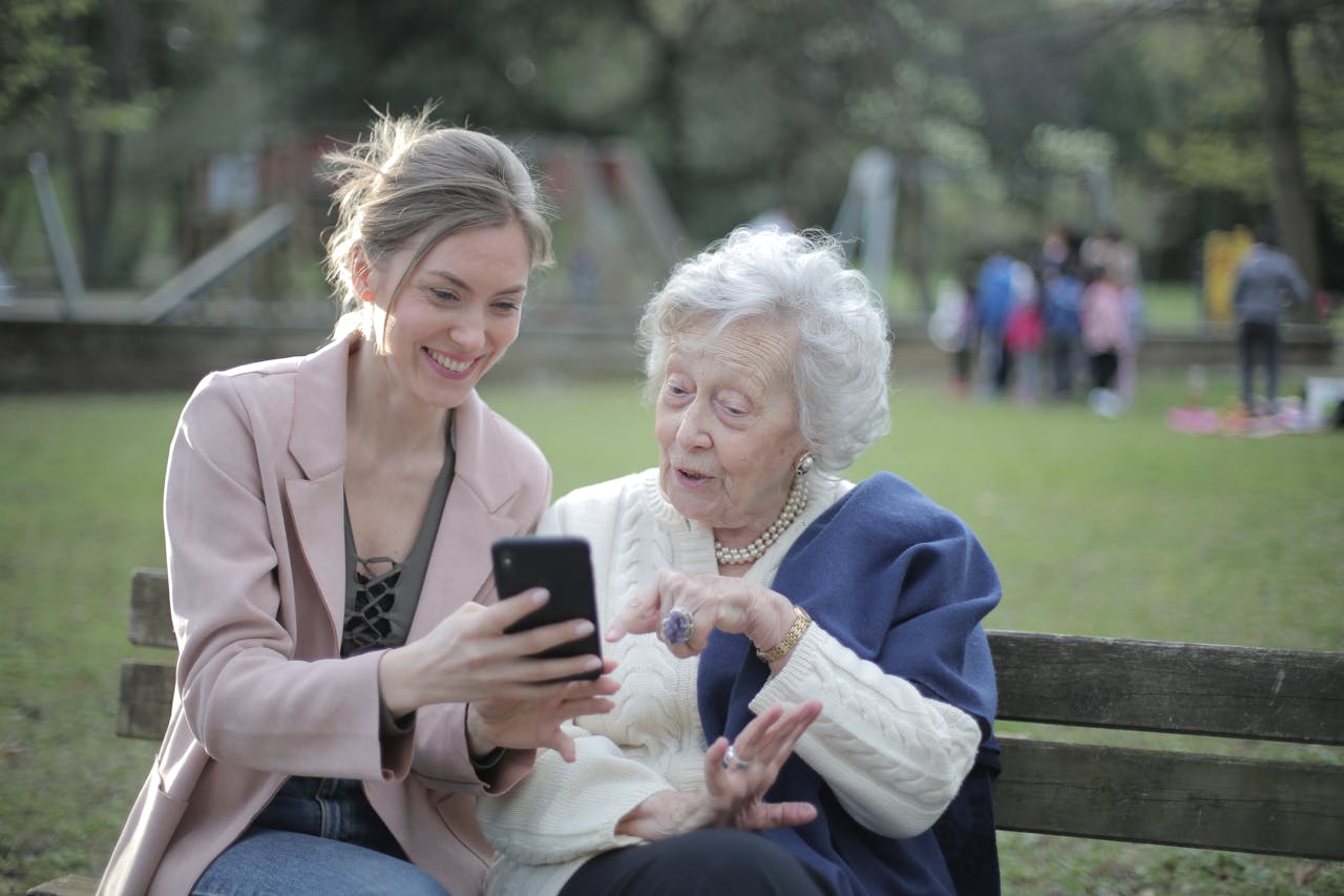 services-02 Delighted female relatives sitting together on wooden bench in park and browsing mobile phone while learning using