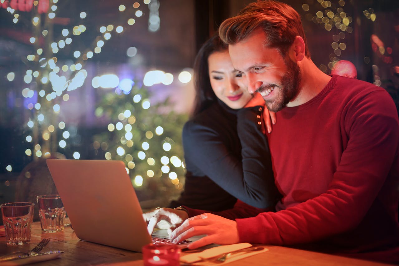 who-we-are A couple using a laptop at a cozy restaurant with festive bokeh lights, enjoying online shopping together.