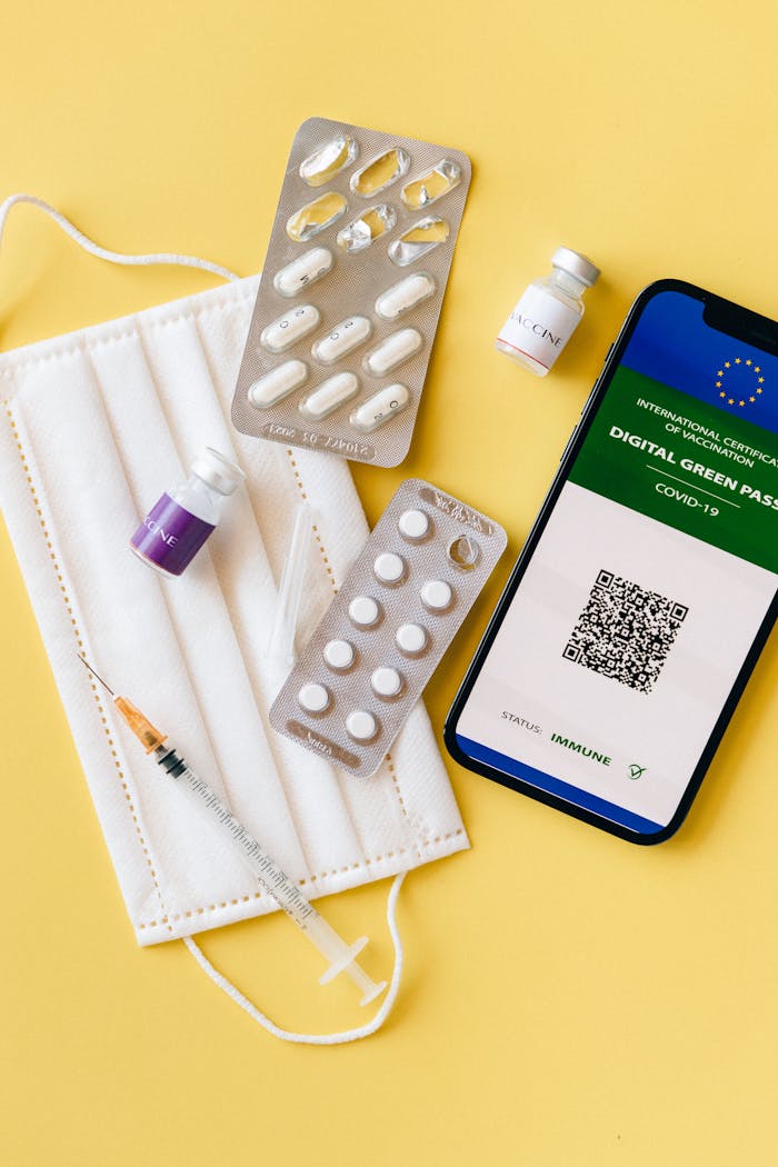services-01 Flat lay of pandemic essentials: mask, vaccine vials, digital pass, medical blister packs, and syringe on a yellow background.